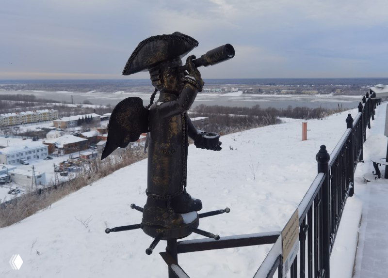 Бронзовая фигурка-ангел на металлическом ограждении смотровой площадки; на заднем плане река и пригородные постройки под зимним небом.