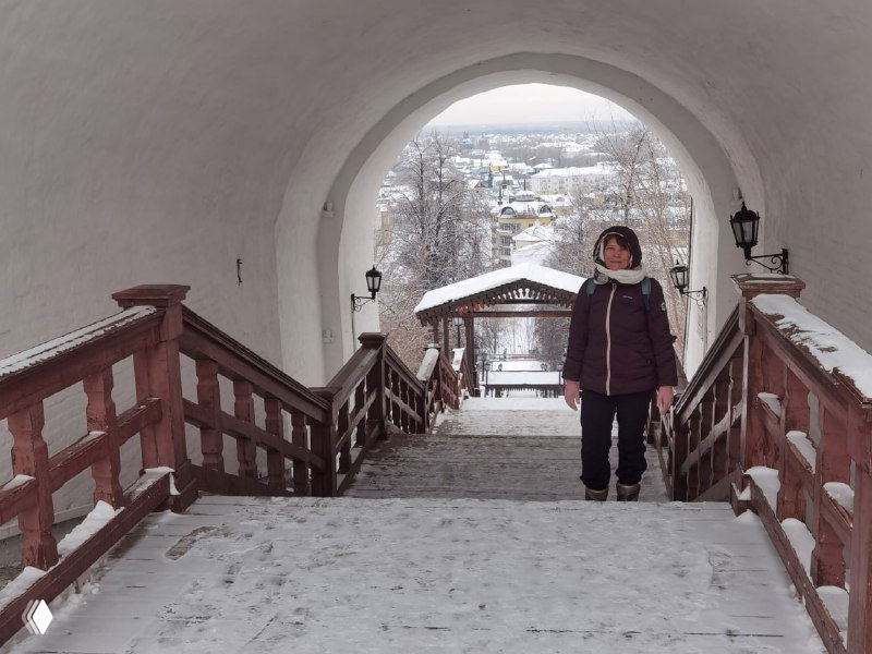 Широкая деревянная лестница, ведущая вниз через арочный проход; туристка в тёплой одежде спускается по ступеням в заснеженном окружении.