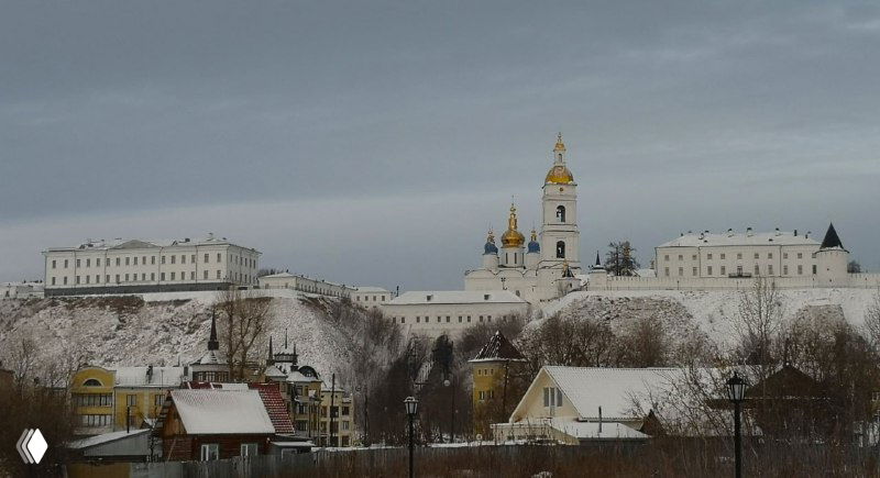 Вид с противоположного берега или возвышенности на белокаменный ансамбль кремля и прилегающие холмы, дома и заснеженные склоны в холодный день.