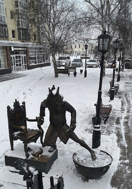 Маленькая скульптурная группа на городской скамейке в садике: фигурки, уличные фонари и заснеженная тротуарная плитка рядом с жилыми домами.