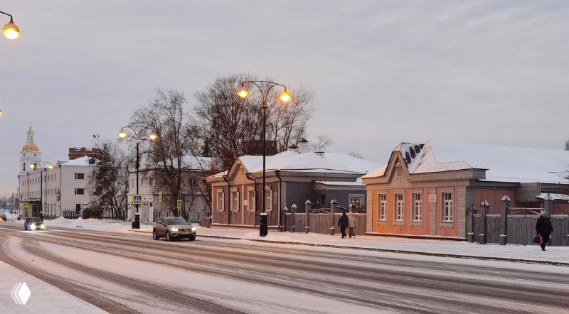 Улица на закате в Тобольске: старинные деревянные дома, вечерние фонари и снежный покров на дороге, создающие атмосферу тихого городского вечера.