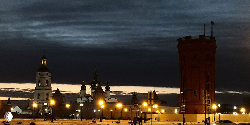 Ночная панорама: освещённые силуэты башен и воды, высокая кирпичная башня и огни города на фоне тёмного неба — вид на кремль в сумерках.