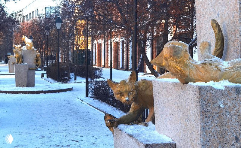 Фотография сквера сибирских кошек в Тюмени: ряд чугунных и бронзовых фигур котов на постаментах, заснеженный городской пейзаж.