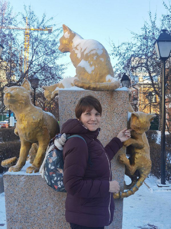 Посетительница у памятника сибирским кошкам в Тюмени: женщина позирует рядом с золотистыми фигурами котов на фоне зимнего сквера.