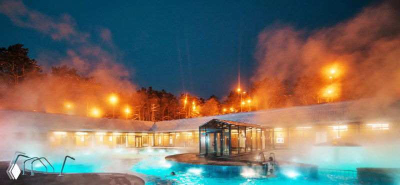 Панорамный кадр открытого термального бассейна вечером: пар, ночные огни и деревянные постройки на фоне соснового леса, атмосфера зимнего отдыха.