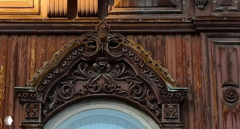 Темно-коричневый фрагмент деревянного фасада с богатой резьбой над арочным окном: растительные и геометрические мотивы в обрамлении.