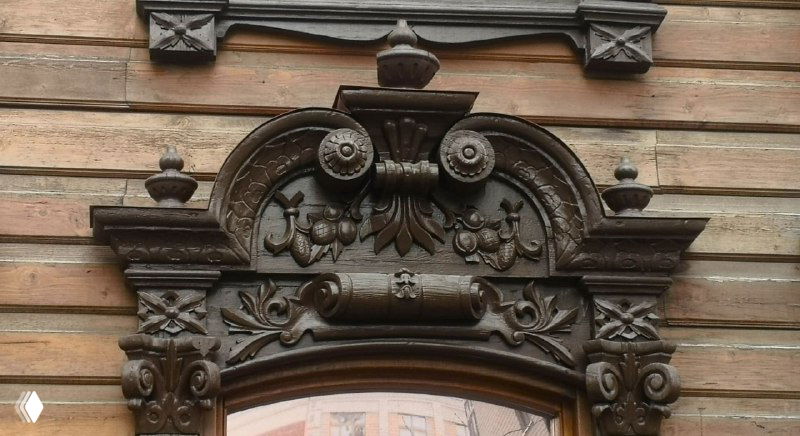 Темная резьба на старом доме с растительными узорами и завитками вокруг окна, выражающая символику и статус владельца.
