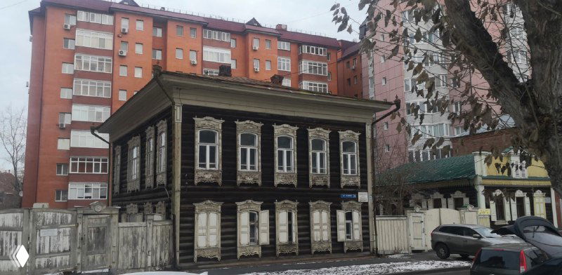 Узкий чёрный деревянный дом на фоне современной застройки в Тюмени — контраст старой городской застройки и новостроек.