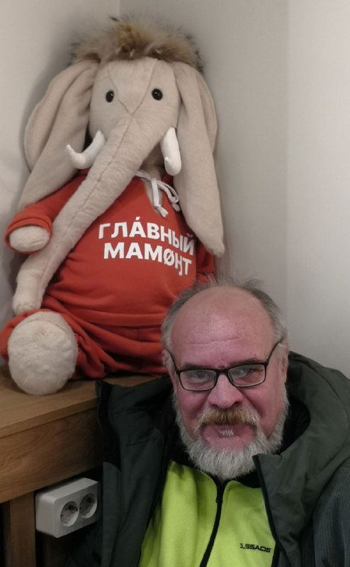 Человек в помещении рядом с игрушечным мамонтом в красной футболке «Главный мамонт», кадр показывает интерьер магазина сувениров или кафе.