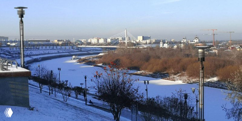 Вид с набережной Туры на зимний городской берег с тропинкой, опорами освещения и мостами вдали, раннее дневное освещение.