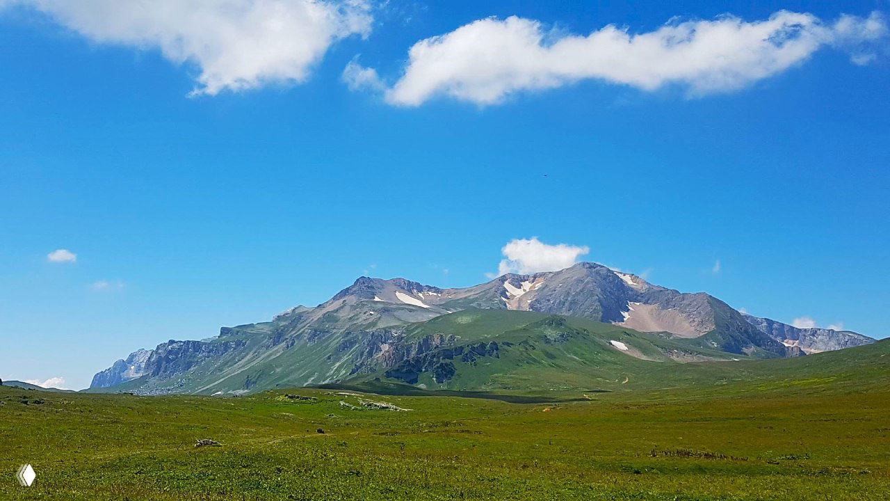 Открытое нагорье и массив Каменное море: широкая ровная терраса перед массивной горой с снежными карманами и обрывистыми скалами на фоне синего неба.