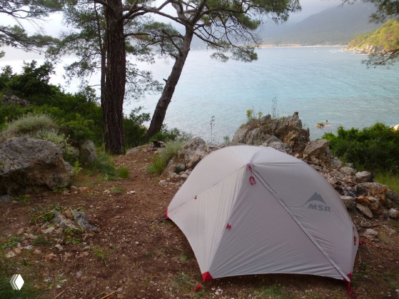 Палатка у скалистого побережья среди сосен с видом на спокойную бухту и море — пример ночёвки в автономном походе по Ликийской тропе.