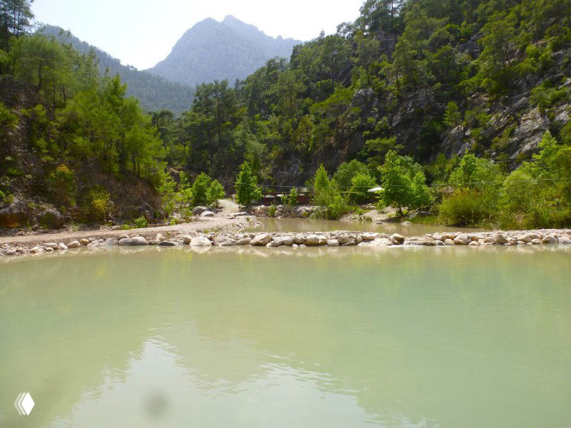 Пологая заводь с мутно-бирюзовой водой и каменистым берегом в нижней части каньона Гёйнюк на фоне зелёных деревьев.