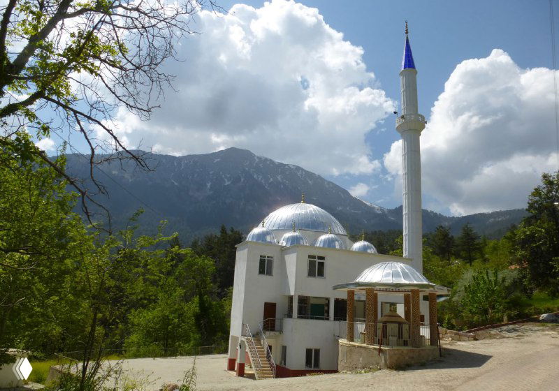 Небольшая мечеть на фоне гор и облачного неба у подножия тропы; вокруг редкие деревья и заросли в предгорье.