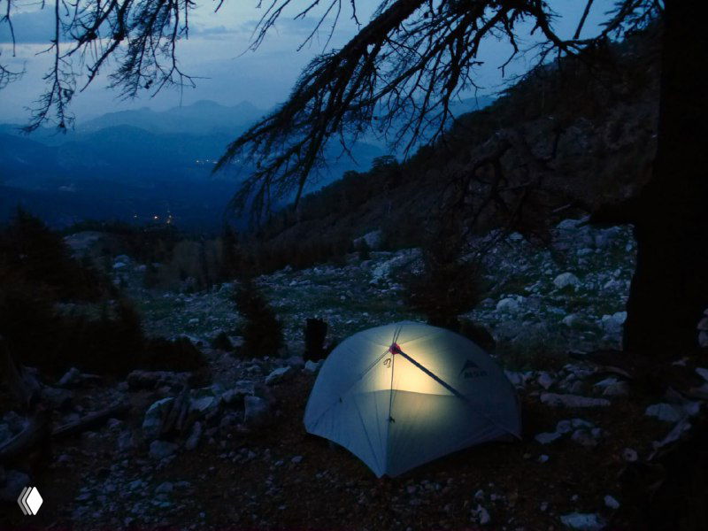 Ночёвка в одиночестве: палатка на каменистой стоянке в сумерках, внутри едва заметный свет и вид на горный склон.