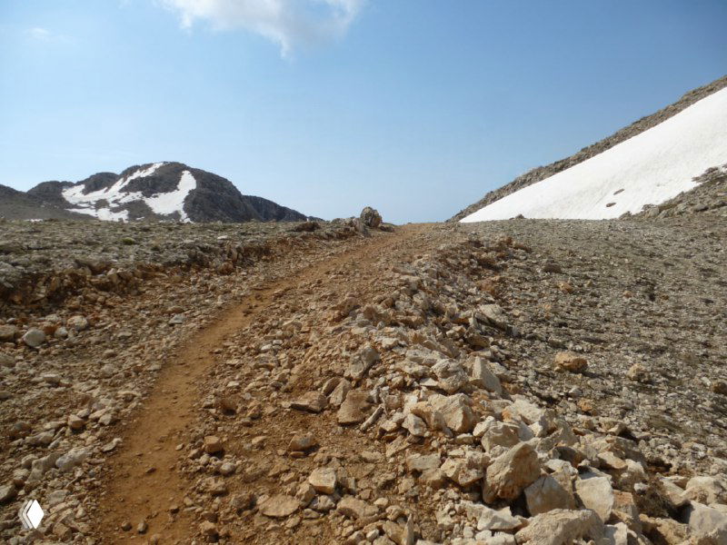 Каменистая горная тропа между островками снега в высокогорье Тахталы — узкая грунтовая дорожка через рыхлые камни и каменные обломки.