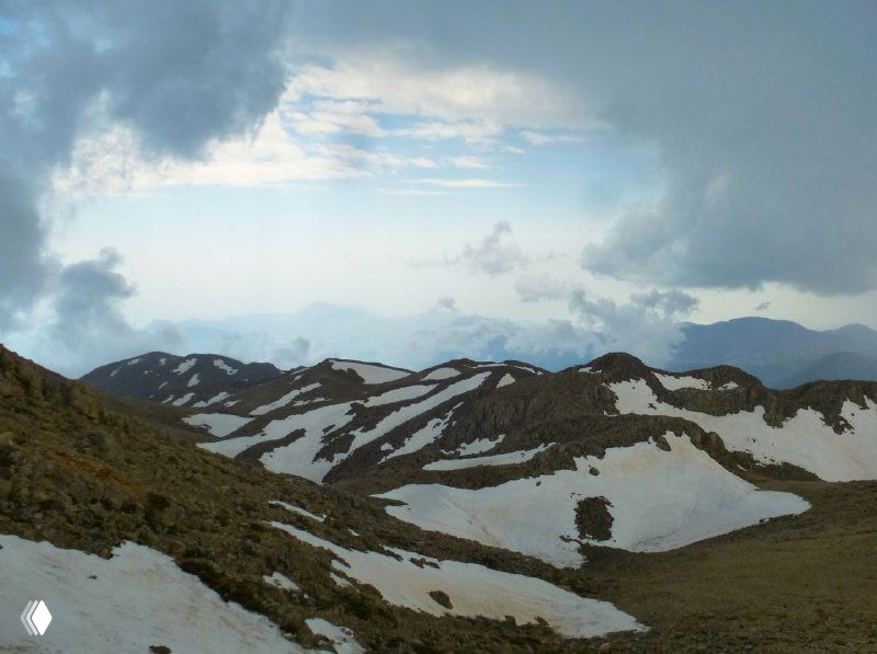 Волнистые горные хребты Тахталы с остатками снега и переменной погодой — широкая перспектива гряд и перевалов в высокогорье.