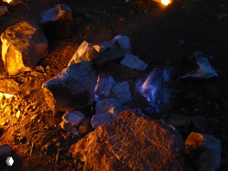 Крупный план синего и оранжевого пламени, вырывающегося между камнями Янарташа; заметна динамика горения природного газа.
