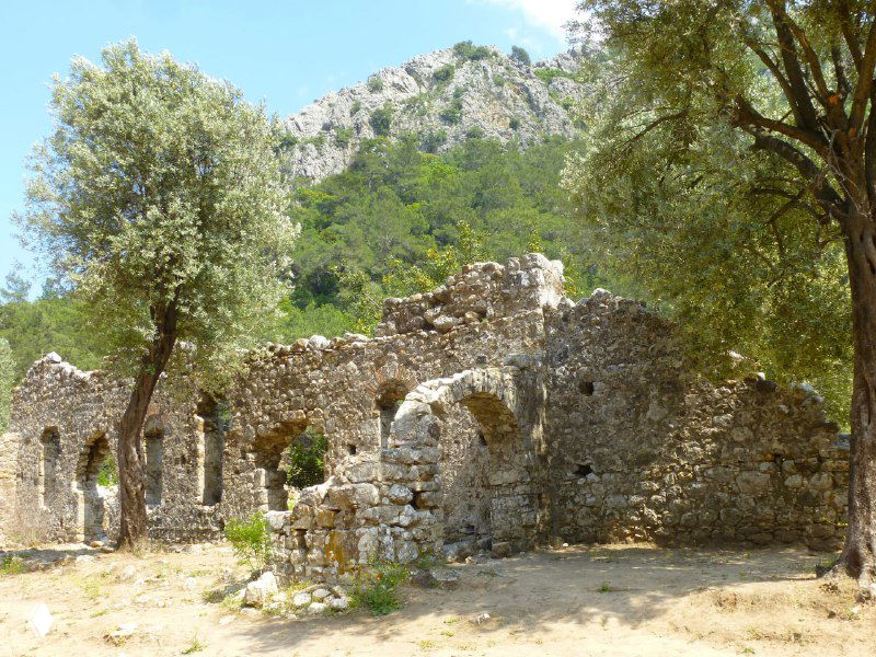 Руины античного Олимпоса среди зелёных деревьев: каменные стены и арки, частично разрушенные и поросшие растительностью.