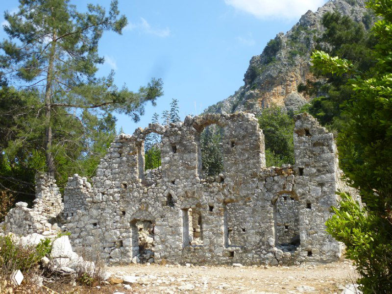 Ещё один ракурс руин Олимпоса: остатки зданий с окнами и стенами под открытым небом, окружённые лесом и холмами.