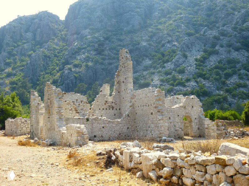 Каменные развалины Олимпоса с хорошо видимыми очертаниями стен и колонн на солнечном открытом участке у подножия скал.