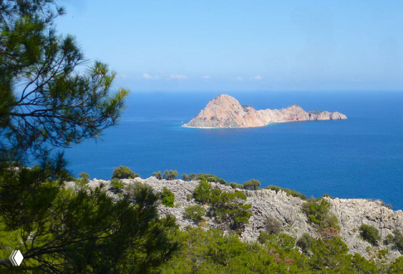 Отдельный скалистый остров в бирюзовом море на подходе к мысу Гелидония, вид с прибрежных склонов Ликийской тропы, подчёркнутый растительностью.