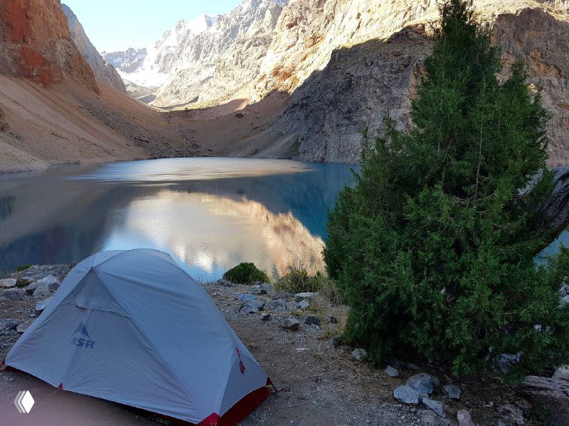 Белая с красным палатка MSR на берегу горного озера среди скалистых стен Фанских гор — спокойный вид лагеря и окружающих хребтов.