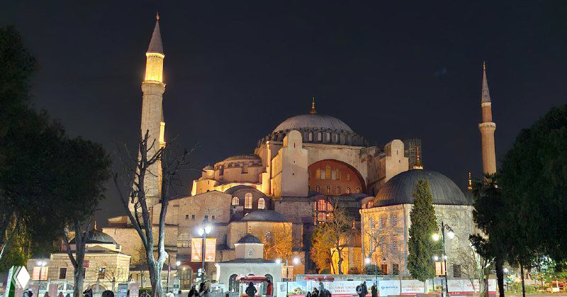 Ночной вид на собор Святой Софии с подсветкой куполов и минаретов, прохожие на площади и тёплое атмосферное освещение исторического центра.