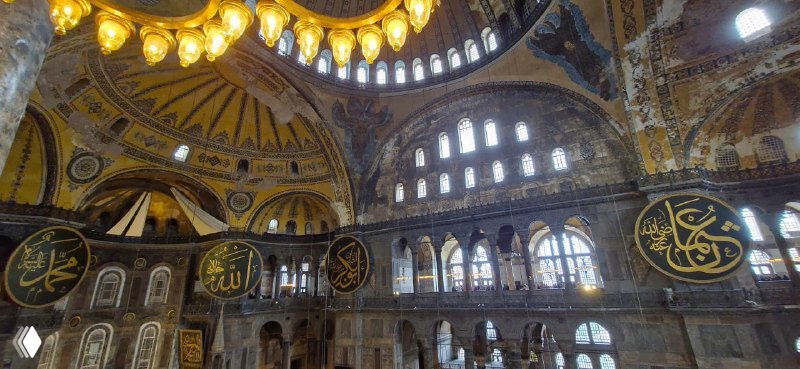 Интерьер Святой Софии с крупными люстрами и круглыми арабскими медальонами на стенах, видны галереи и высокие окна, пропускающие рассеянный свет.