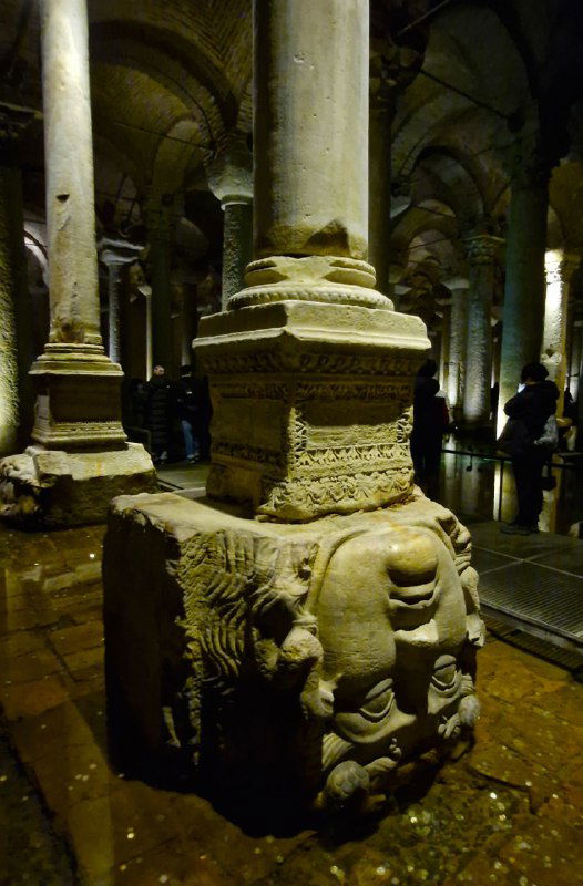 Крупный план одной из колонн с перевёрнутой головой Медузы в основании: каменная скульптура, частично погружённая в воду, с текстурами времени.