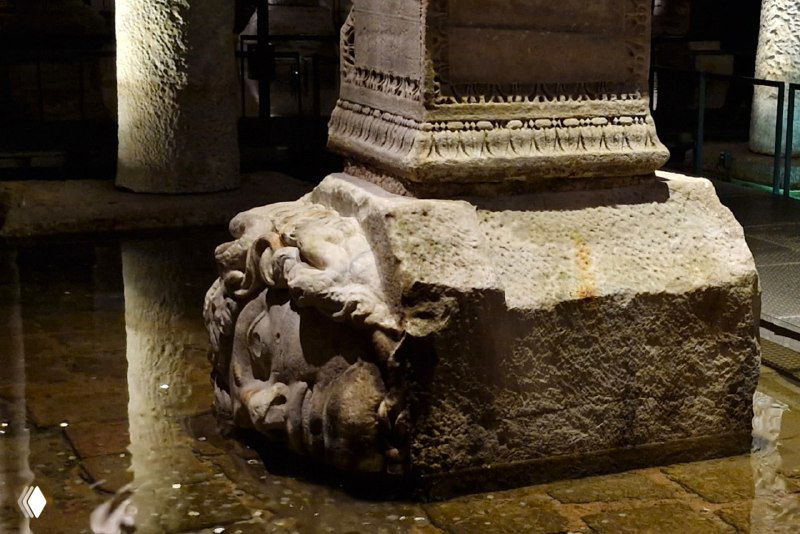 Крупный план каменного блока с барельефом и очертаниями головы, стоящий на мокром полу цистерны, демонстрирующий детализацию античных остатков.