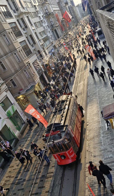 Вид с высокого ракурса на улицу Истикляль: красный трамвай движется по рельсам среди пешеходов, витрин и уличных лавок в плотной толпе.