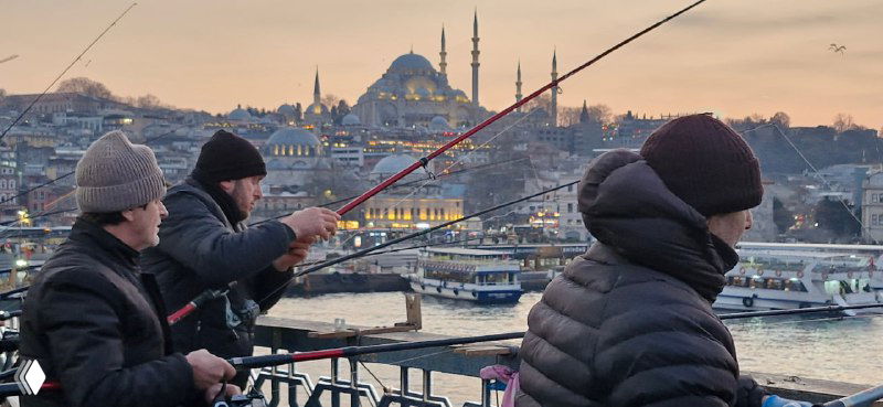 Рыбаки на Галатском мосту забрасывают удочки на фоне закатного неба и силуэта мечетей Стамбула, люди любуются рыбалкой с моста.