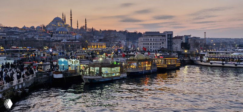 Прибрежная сцена у Золотого Рога: лодки, прилавки с балык экмек и люди на пирсе на фоне городской набережной и заката.