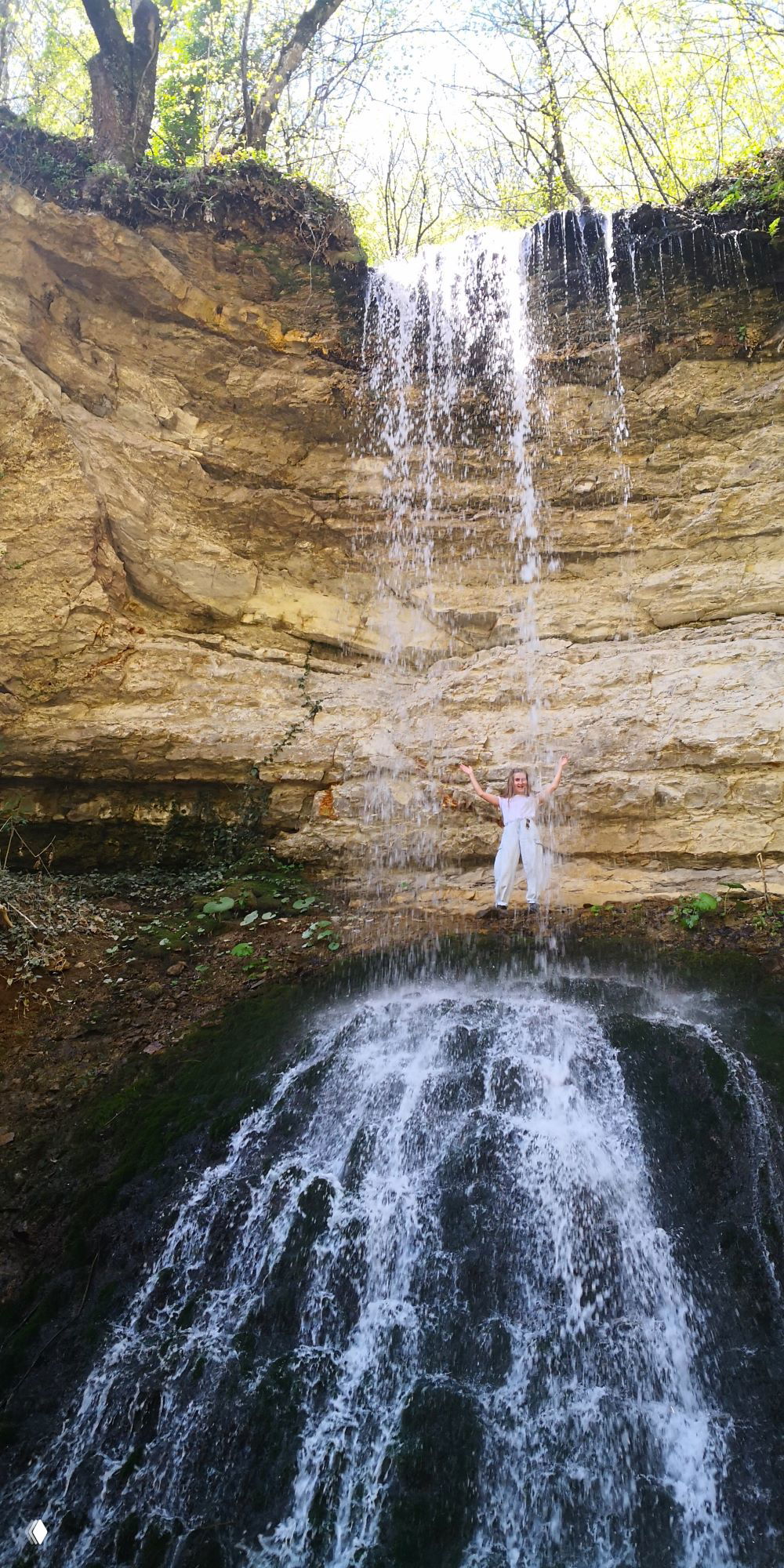 Тонкая струя водопада падает с карниза скалы, на фоне виден человек за струёй, вокруг светлые деревья и известняковые уступы.