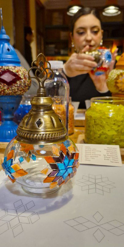 Готовая маленькая мозаичная лампа на столе музеобразного мастерского помещения, видно сочетание голубых и оранжевых стеклышек и металлический подвес.