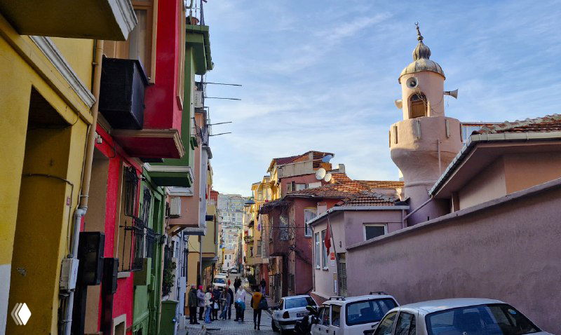 Узкая улица Фенера с разноцветными домами, людьми и минаретом или куполом на горизонте в ясный день