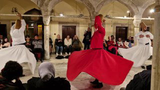 Кружащиеся дервиши в широких юбках во внутреннем зале с арочными колоннами; зрители у стен наблюдают за вечерним шоу.