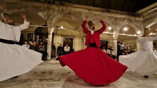 Красный танцор-дервиш в развевающейся юбке на фоне белых дервишей и архитектуры исторического зала; зрители по периметру.