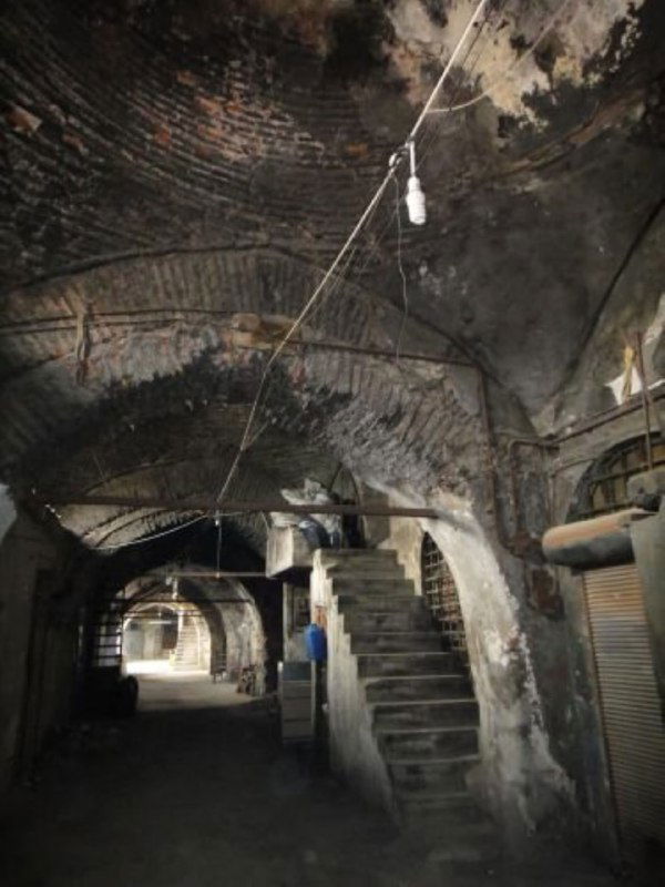 Тёмный узкий коридор внутри Валиде Хан с арочными сводами, лестницей и атмосфёрной полуосвещённостью — интерьер караван-сарая в запустении.