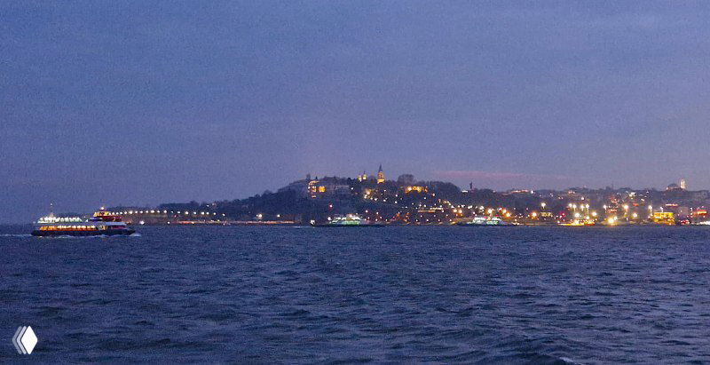 Ночной вид бухты и городского берега Стамбула с подсвеченными домами и проплывающим паромом по тёмному Босфору на дальнем плане.