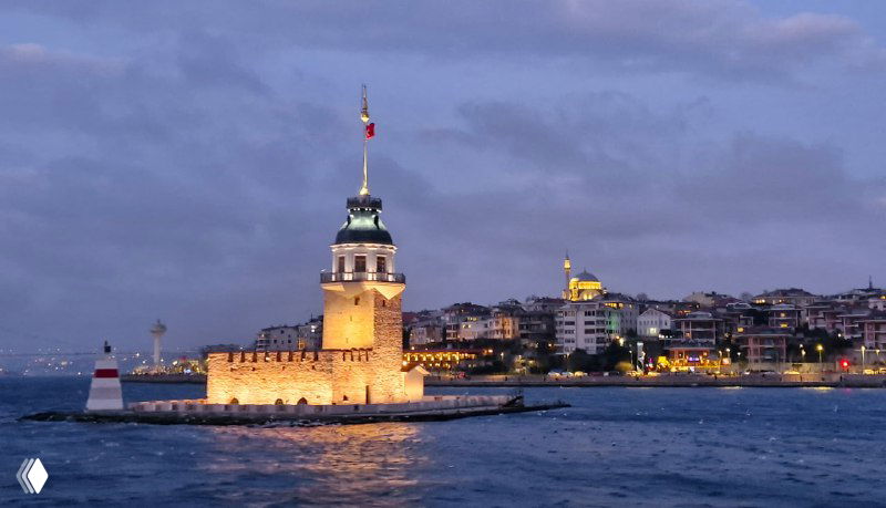 Близкий план крепости Девичья башня (Kız Kulesi) на маленьком островке, вечерняя подсветка и здания азиатского берега за ней.