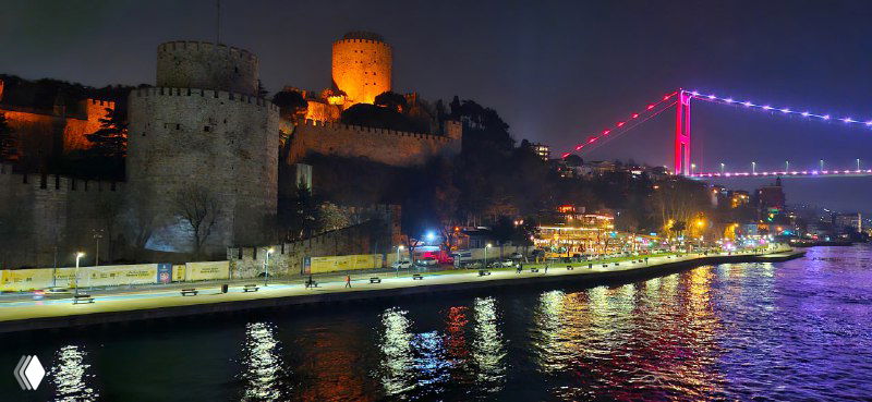 Вид крепостной стены и прибрежных построек вдоль Босфора, тёплые огни архитектуры и освещённый мост вдалеке, отражающийся в воде.