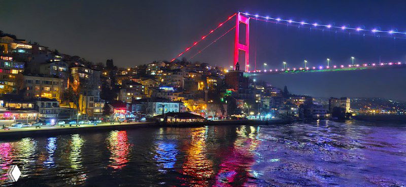 Подсвеченный ночью мост через Босфор, разноцветная иллюминация и отражение в воде, лайнеры и огни вдоль береговой линии.