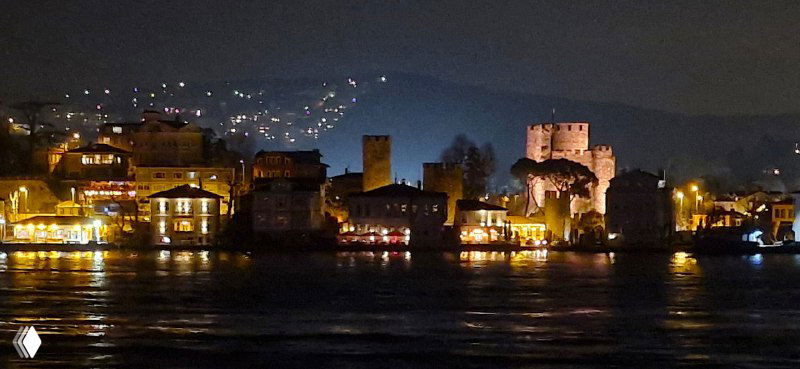 Ночной прибрежный пейзаж с крепостями и домами у воды, тёплые огни вдоль набережной и спокойная поверхность Босфора с лёгкими рябью.