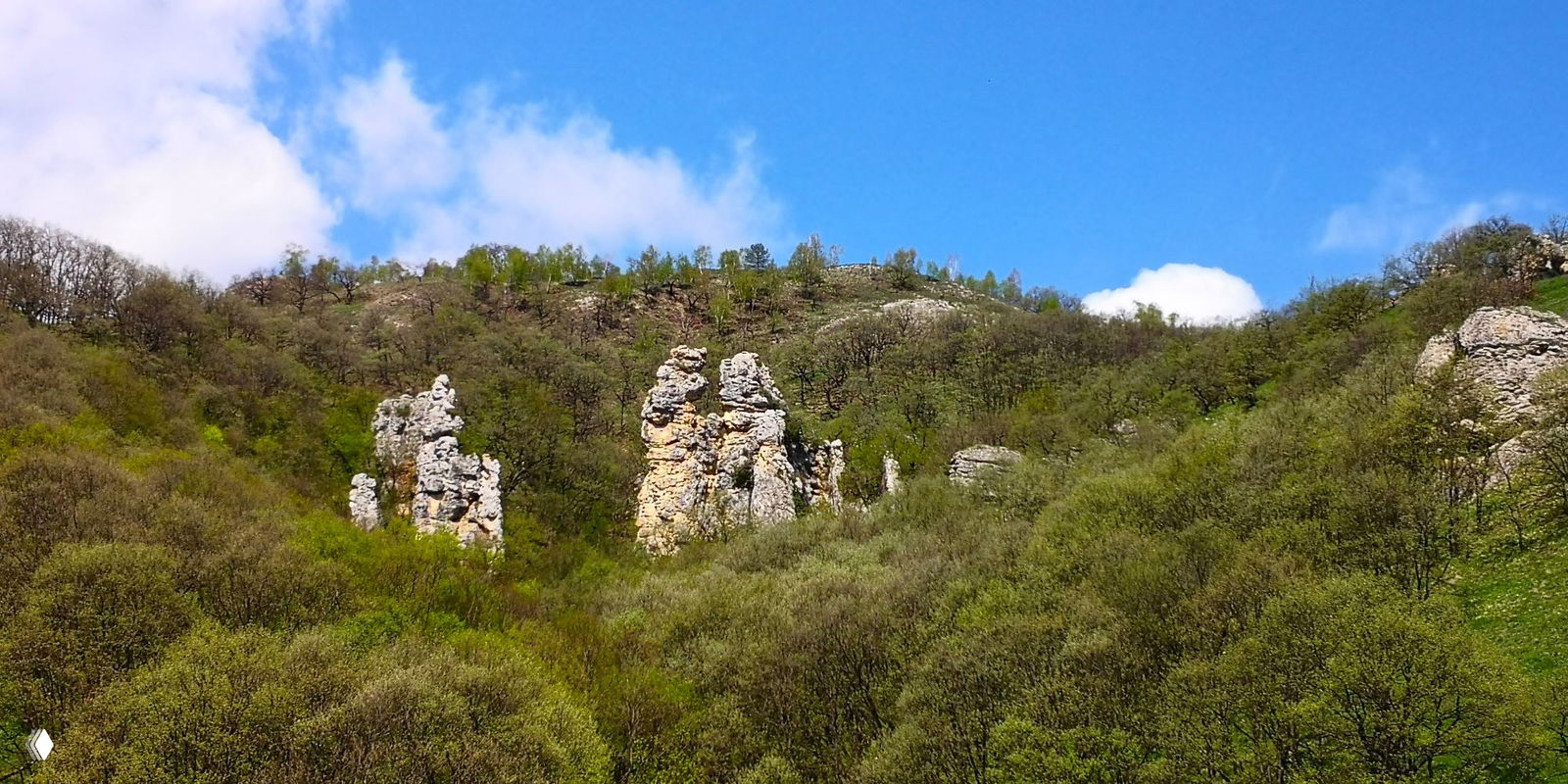 Малый водопад у скального выступа: турист стоит рядом и тянет руки к падающим струям, внизу каменистое русло и корни деревьев.