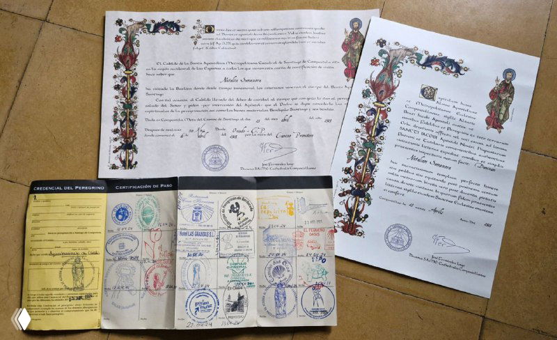Креденсиаль рядом с оформленной компостелой и сертификатами: листы с латинским текстом и печатями, документ и паспорт пилигрима на фоне пола.