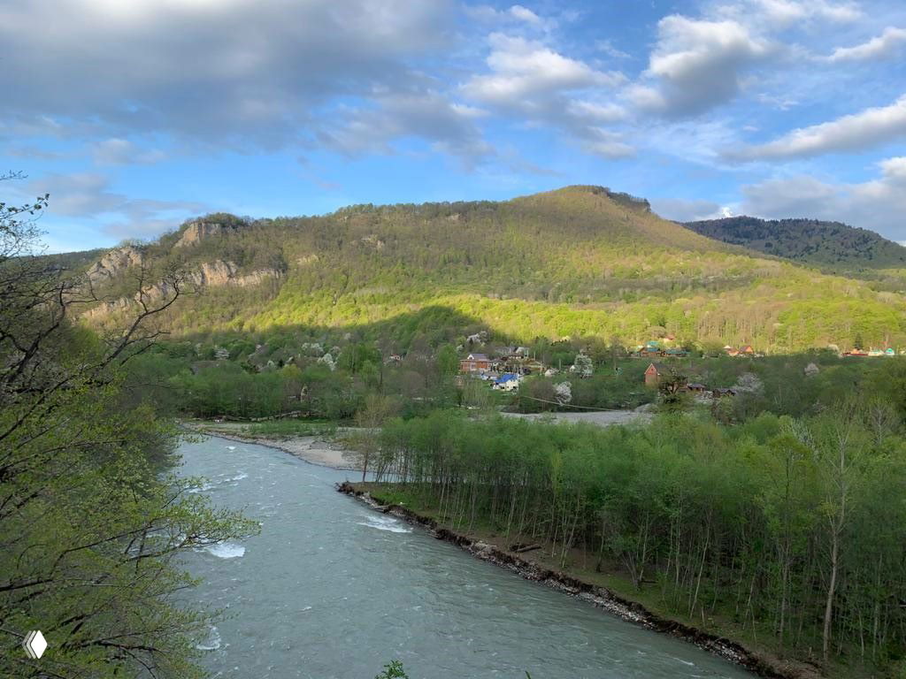 Речная долина и зеленые склоны, спокойный участок Малой Лабы и дома на дальнем берегу в ясный день с облачным небом.