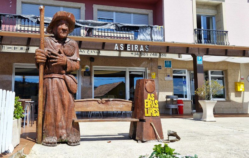 Деревянная скульптура паломника с посохом на площади у входа в альберге и магазинчики на фоне — местный колорит маршрута Камино.