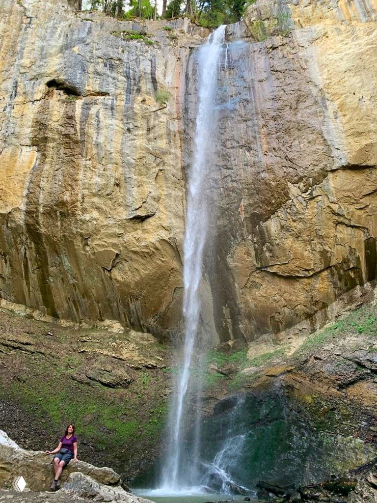 Высокий тонкий водопад, стекающий по светло-коричневой отвесной скале в узкую чашу у её основания, вид снизу.